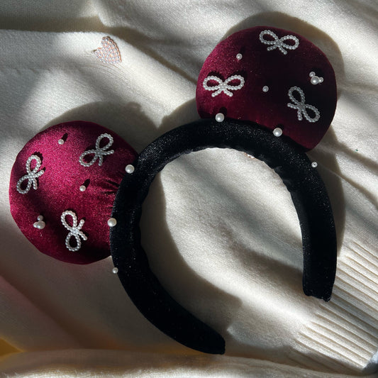 A close-up of a headband with wine red velvet ears and a black velvet headband, adorned with pearl bows and mouse head accents.